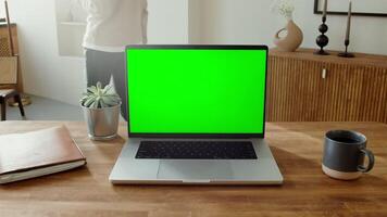 Close-up of an open laptop with a green screen standing on a table in a home interior. A woman is walking in the background, her face is not visible. Copy space. in 4k format, red komodo. video