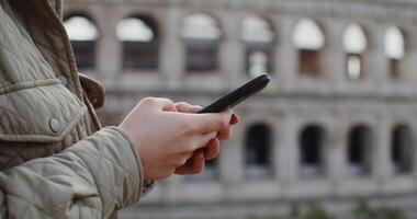 A young warmly dressed woman dials a message on her mobile phone while standing in front of the Coliseum in Rome. Women's hands close-up, the face is not visible. in 4k, red komodo. video