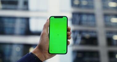 A man's hand in a business suit holds a mobile phone with a green screen on the background of a modern building. Copy space, mockup, chroma key. in 4k, red komodo video