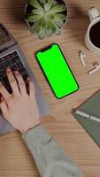 A mobile phone with a green screen lies on the desktop next to female hands typing on a laptop, vertical , close-up. Chroma key, mockup, copy space. in 4k format, red komodo. video