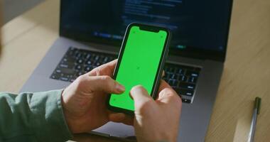 A young fair-skinned man is typing on a green screen of a mobile phone, interrupted from work at a laptop. Filming from behind, the man's face is not visible, chroma key, copy space video