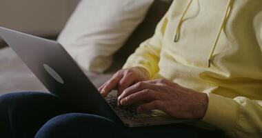 An elderly man of European appearance, dressed in casual clothes, uses a laptop while sitting on the couch at home, close-up of male hands typing on the keyboard, the man's face is not visible video