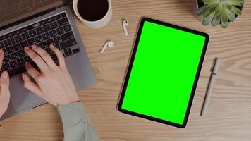 Close-up of a digital tablet with a green screen lying on a table next to female hands typing on a laptop. Chroma key, mockup, copy space. in 4k format, red komodo. video