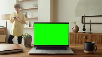 Young woman typing text on mobile phone standing behind open laptop with green screen standing on table, copy space, chromakey, mockup. in 4k format, red komodo. video