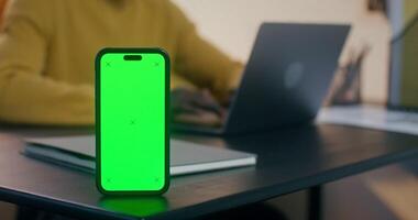 A mobile phone with green screen is on the desktop. Men's hands typing on a laptop in the background video