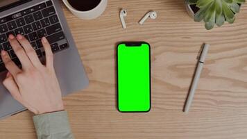 A mobile phone with a green screen lies on the desktop next to a laptop on which a woman is typing, a close-up of female hands. Chroma key, mockup, copy space. in 4k format, red komodo. video