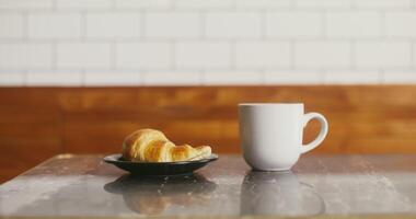A large white mug and a black plate with a croissant stand on a small coffee table, prepared for a regular customer. No people on video