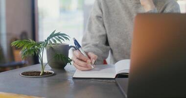 Young woman dressed in casual clothes making notes with a pen in a notebook sitting at a table in front of an open laptop, close-up, the woman's face is not visible video