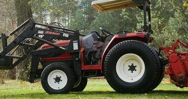 A modern agricultural tractor with a canopy above the driver's seat, with a bucket in front, stands at the edge of a field near the forest, close-up video
