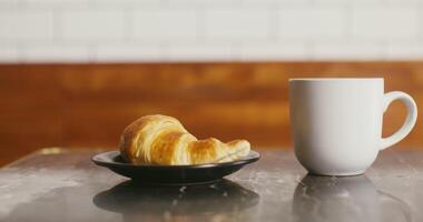 An appetizing croissant on a plate and a mug of coffee are on a small table ready for breakfast. No people on video
