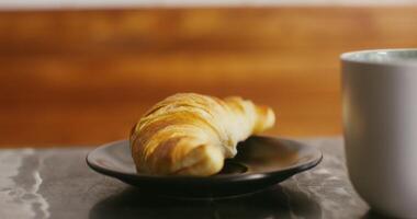 An appetizing fresh croissant lies on a small black plate on the table next to a coffee mug, close-up video