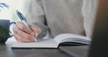 Woman's hand close up. A young woman makes notes by a pen in a notebook sitting at a table in front of an open laptop. A pot with a houseplant is built into the table. The woman's face is not visible video