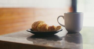 A large white mug with steaming hot coffee and a black plate with a croissant stand on a small table in a cafe, ready for lunch video