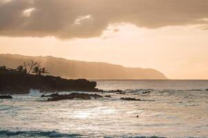 A serene coastal scene at sunset on Oahu, Hawaii, featuring gentle waves, a lone swimmer, silhouetted palm trees, and distant mountain ridges. photo