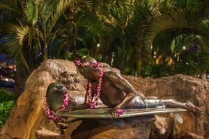 A bronze statue of a surfer on a surfboard, adorned with Hawaiian flower leis, set against tropical plants and rocks under soft nighttime lighting. photo