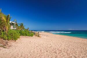 A tranquil beach with clear turquoise waters, a wooden fence pathway, lush green vegetation, palm trees, and a small structure on the left. photo