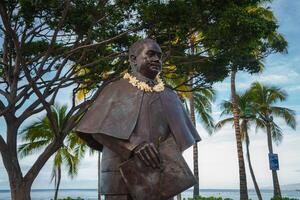 A bronze statue of a man with a traditional lei stands near palm trees, lush greenery, and a coastal view under a clear blue sky in Hawaii. photo