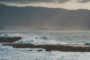 Waves crash against rocky outcrops along a tropical coastline, with mist covered mountains and sunlight breaking through a cloudy sky in the background. photo