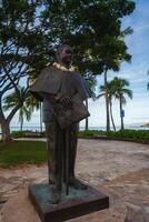 A bronze statue of a man with a lei stands on a stone pedestal in a park with palm trees, benches, and an ocean view, surrounded by tropical greenery. photo