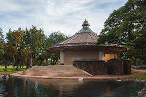 A circular pavilion with a conical roof, stone steps, and columns, surrounded by lush greenery, tall trees, and a serene pond in a Hawaii park. photo