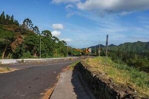 A scenic road bordered by greenery and a stone wall curves left, with hills and mountains in the background under a clear sky with scattered clouds. photo