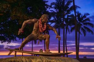 A bronze surfer statue adorned with flower leis stands on a surfboard, framed by palm trees, a vibrant sunset, and a serene Hawaiian beach. photo