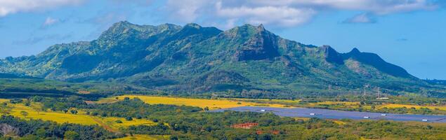 Aerial view of Kauai Island, Hawaii, showing green mountains, rugged ridges, fields with mixed vegetation, and a solar farm under a partly cloudy sky. photo