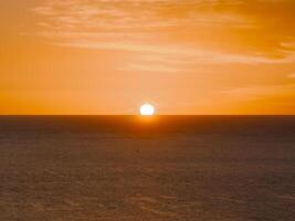 The sun sets at the horizon, casting an orange glow over the ocean. A small boat silhouette is visible in the tranquil expanse of water and sky. photo