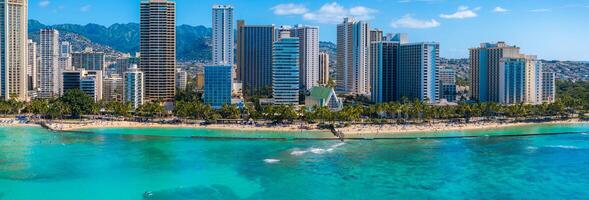 Turquoise waters meet sandy shores at Waikiki Beach, with palm trees, beachgoers, modern skyscrapers, and lush green mountains in the background. photo