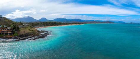 Aerial view of Oahu island's coastline featuring turquoise waters, a sandy beach, rocky outcrops, lush greenery, and a mountain range under a cloudy sky. photo