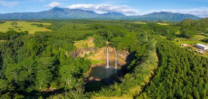Twin waterfalls cascade into a tranquil pool, surrounded by lush vegetation, dense forests, open fields, and dramatic cloud covered mountains. photo