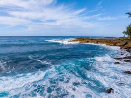 Vibrant blue ocean waves crash against a rocky shoreline on Oahu, Hawaii, under a partly cloudy sky, with trees and vegetation along the coast. photo