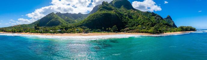 Aerial view of a tropical beach on Kauai island, Hawaii, with golden sand, turquoise waves, lush Napali cliffs, palm trees, and small structures. photo
