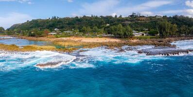 Turquoise waves crash against a rocky shoreline in Oahu, Hawaii. A sandy beach is bordered by greenery, with homes and trees in the background. photo