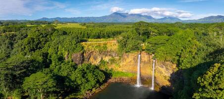 Twin waterfalls cascade over a green cliff into a pool, surrounded by tropical vegetation, with fields and mountains visible in the background. photo