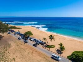 Aerial view of a beach on Oahu island, Hawaii, featuring golden sand, turquoise ocean, a narrow road with parked cars, palm trees, and clear skies. photo