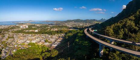 Aerial view of Oahu, Hawaii, showing a highway through green hills, a residential area, rugged mountains, and a distant coastline under bright daylight. photo