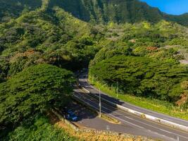 A winding road bordered by dense tropical trees at the base of a forested mountain on Oahu island, Hawaii, with cars parked along the roadside. photo
