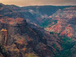 View of Waimea Canyon on Kauai Island, Hawaii, featuring steep cliffs with red, brown, and green layers, contrasting with lush vegetation below. photo