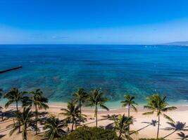 Aerial view of a beach on Oahu island, Hawaii, with turquoise waters, coral reefs, a small pier, palm trees, and boats under a clear blue sky. photo