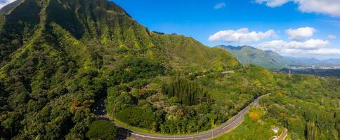 Aerial view of green mountains and valleys on Oahu Island, Hawaii, with a winding highway cutting through dense tropical vegetation under a clear sky. photo