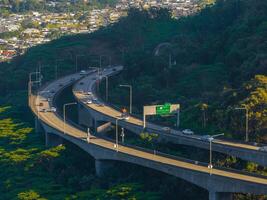 An elevated highway curves through dense vegetation on Oahu island, Hawaii, with a residential area in the background and a green road sign visible. photo