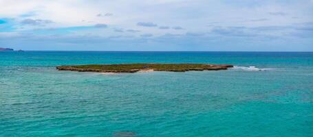 A flat island with low vegetation and a sandy area surrounded by calm turquoise waters. People are visible, with blue skies and scattered clouds above. photo
