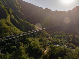 Highway bridge spans green mountains on Oahu Island, Hawaii, with sunlight casting warm tones and rugged ridges in the background. photo