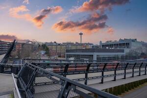 View of Seattle with the Space Needle, a modern pedestrian bridge, colorful buildings, and greenery under a vibrant sunset sky with scattered clouds. photo