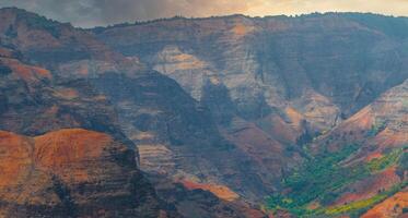 Colorful cliffs of Waimea Canyon on Kauai Island, Hawaii, with red and orange rock layers, green vegetation, and a cloudy sky creating a dramatic scene. photo