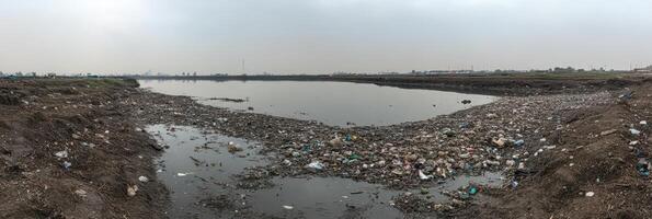 Polluted riverbank covered with trash and debris in urban environment under cloudy sky photo