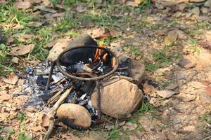 Classic three legged iron stand is placed over an open fire, designed to hold cooking pots. This method reflects traditional outdoor cooking, commonly used in camping and rural kitchens. photo