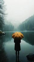 Person with yellow umbrella stands by tranquil lake in misty forest during early morning. photo