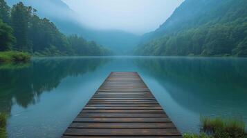 Tranquil lake surrounded by misty mountains at dawn with a wooden pier extending into the water. photo